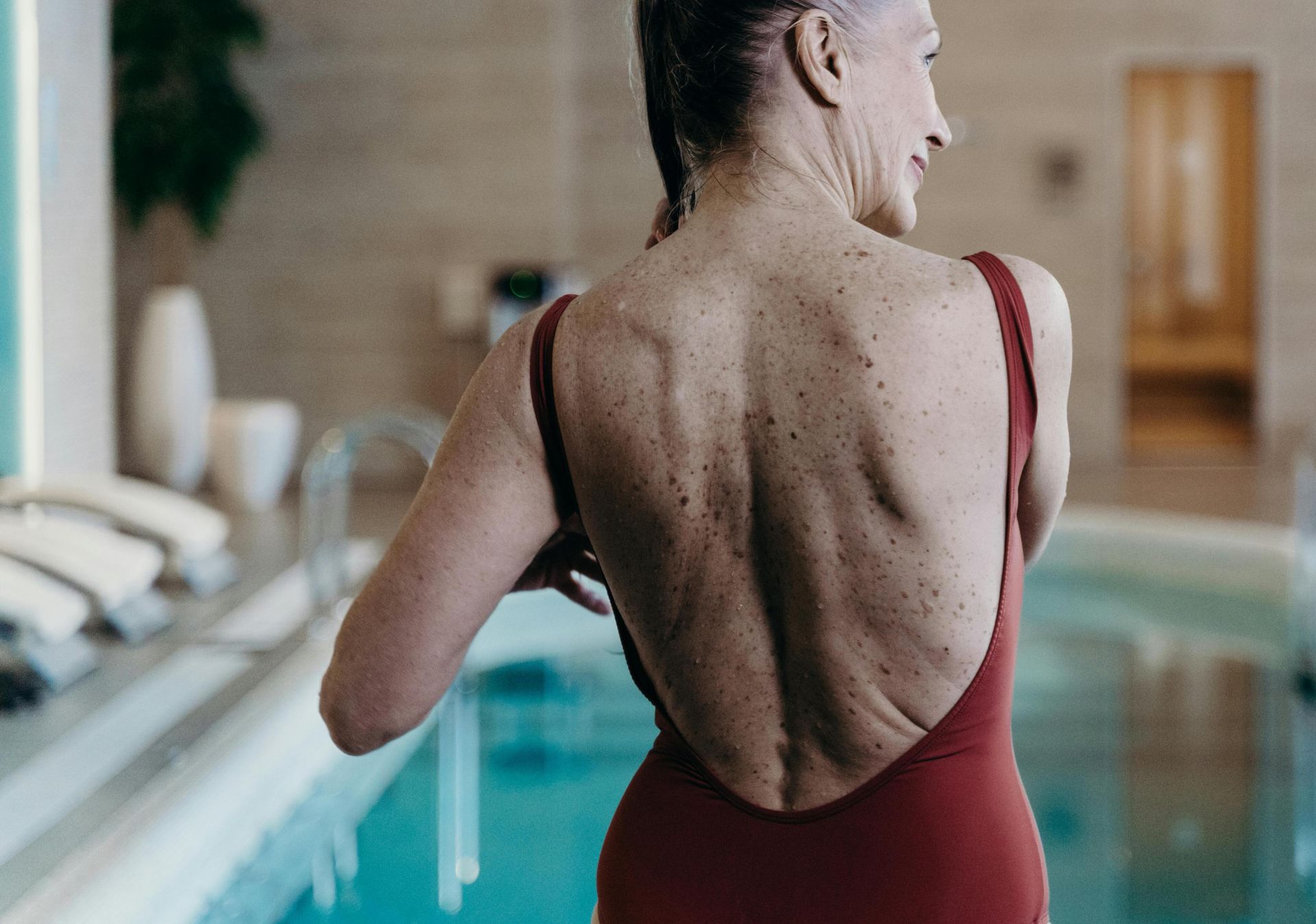 Senior woman in red swimsuit standing at indoor pool side, exuding confidence.