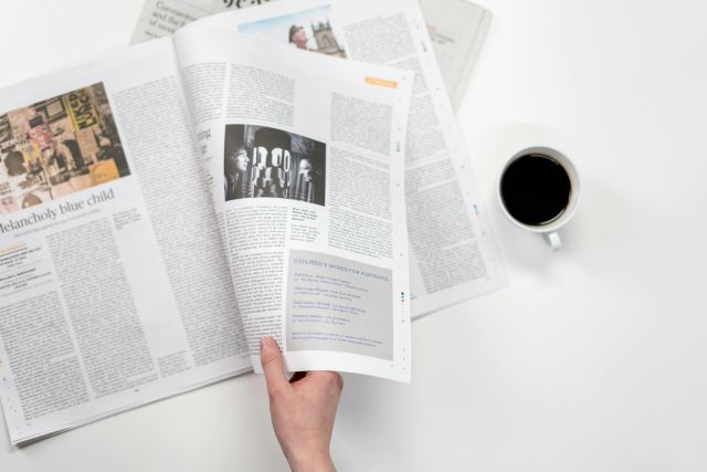 A close-up shot of a hand holding a newspaper with a coffee cup on a white surface.